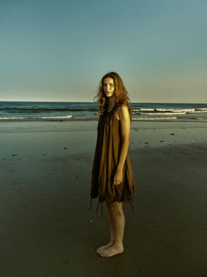 Model standing on a beach at dusk wearing a flowing brown dress